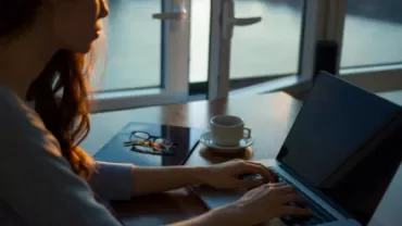 Woman typing on a laptop with a cup of coffee on a wooden table.