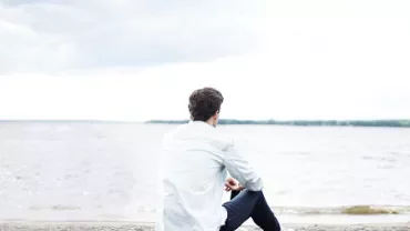 Person sitting by a beach looking at the water under a cloudy sky.