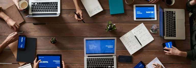 Top view of people working on laptops and tablets at a wooden table.