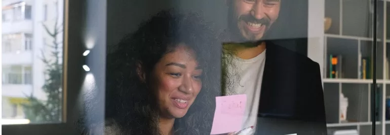 Two people smiling and reading sticky notes on a glass wall in an office.