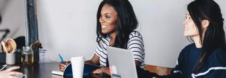 Two women at a table with laptops and coffee, engaged in discussion.
