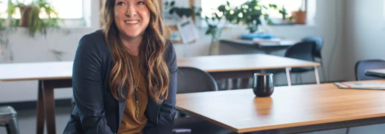 Smiling person sitting in a modern office with wooden tables and plants.