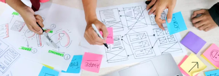 People brainstorming at a desk with wireframe sketches and colorful sticky notes.