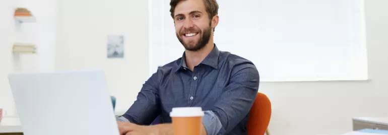 Smiling man in a blue shirt working on a laptop at a desk with a coffee cup.