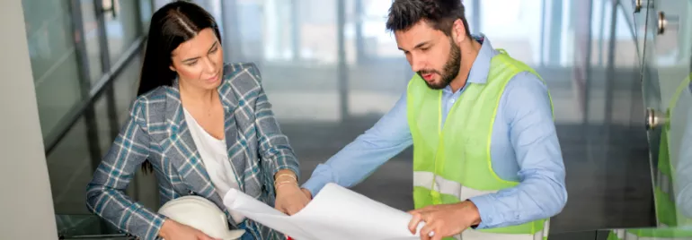 Two professionals reviewing building plans in an office corridor.
