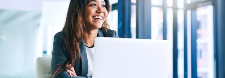 Woman in a suit smiling while working on a laptop in a bright office.