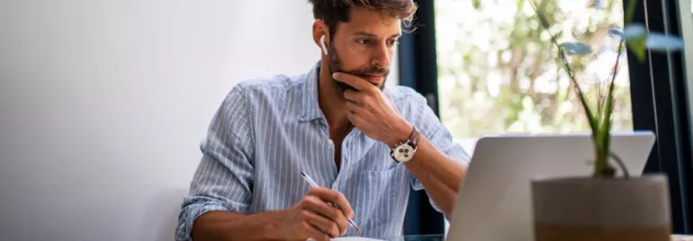 Man wearing earphones working on a laptop while taking notes.