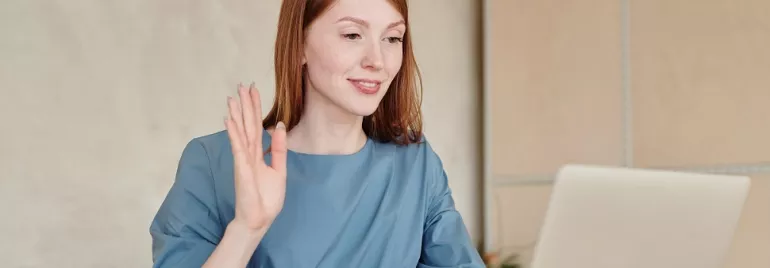 Woman in blue shirt waving at laptop during video call.