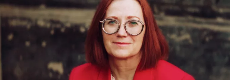 Woman with red hair and glasses wearing a red jacket.