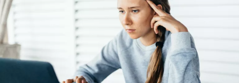Young woman focuses intently on laptop screen, holding pen.
