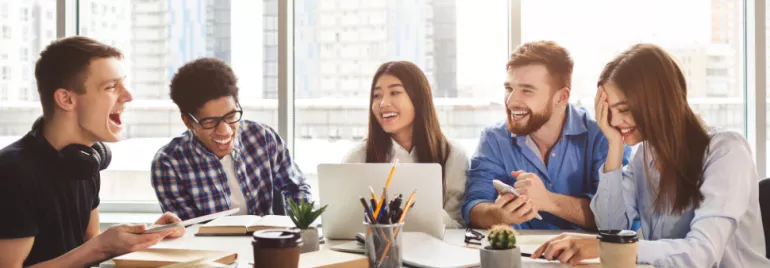 Group of five young adults laughing and working around a table with notebooks and laptops.