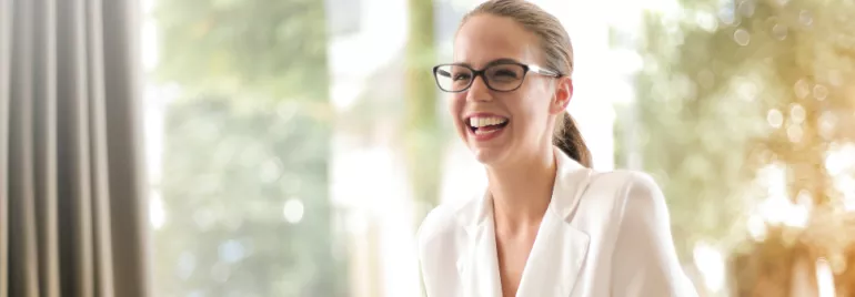 Woman in glasses laughing while sitting in a bright room.