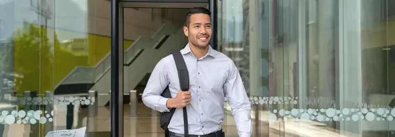 Smiling man with a backpack exits a glass building entrance.