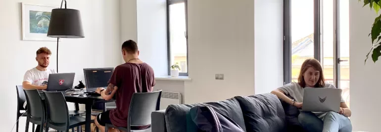 Three people working on laptops in a bright room with large windows.