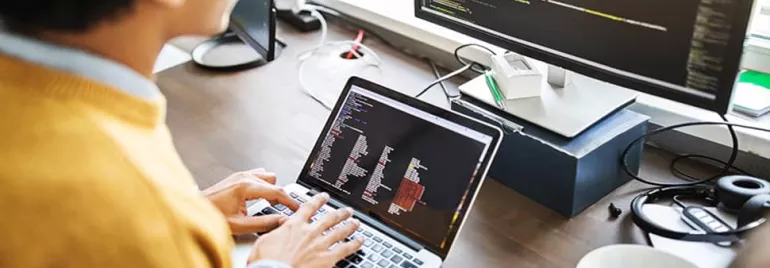 Person coding on a laptop with a larger screen displaying code.