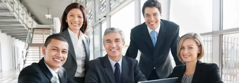 A diverse group of business professionals smiling around a laptop in a modern office.