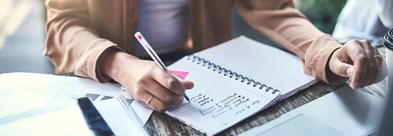 Person writing notes in a spiral notebook at a desk with documents and a laptop.
