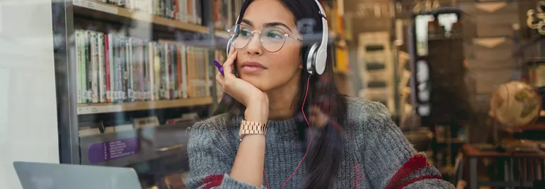 Woman wearing headphones looking out a library window, with books in the background.