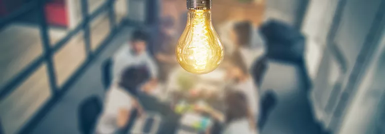 Close-up of a lit light bulb with blurred people working in the background.