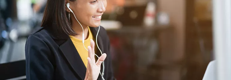 Woman smiling and waving during a video call, wearing earphones, with a blurred background.