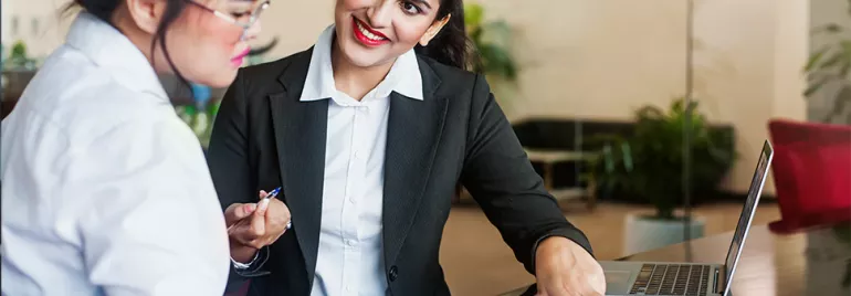 Two women having a discussion at a desk in an office setting.
