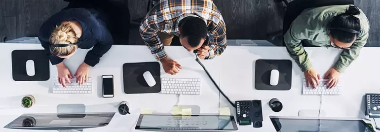 Overhead view of three people working at office desks with computers, using headsets and keyboards.