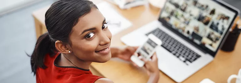 Woman smiling while holding phone at desk with laptop open.