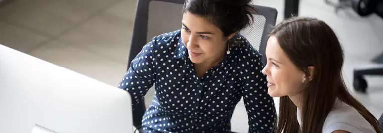 Two women smiling and working together at a computer desk in an office setting.