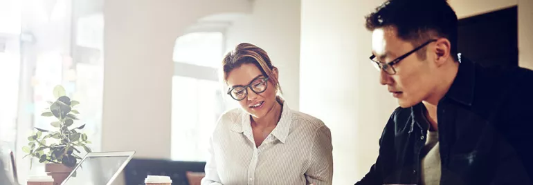 Two people working together on documents at a table with coffee and a laptop.