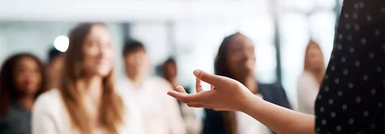 A speaker gesturing in front of a blurred audience.
