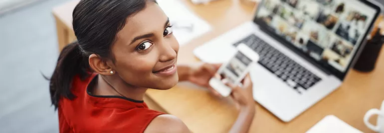 Woman in red top smiling, holding a phone, looking at a laptop with images.