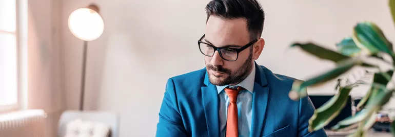 Man in blue suit and glasses working on a laptop in a modern office.