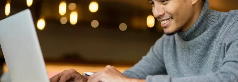 Man in gray sweater smiling while using a laptop.