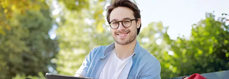 Smiling person with glasses sitting outdoors, holding a tablet.