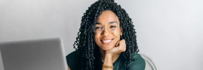 Woman with glasses smiling at laptop, hand on chin.
