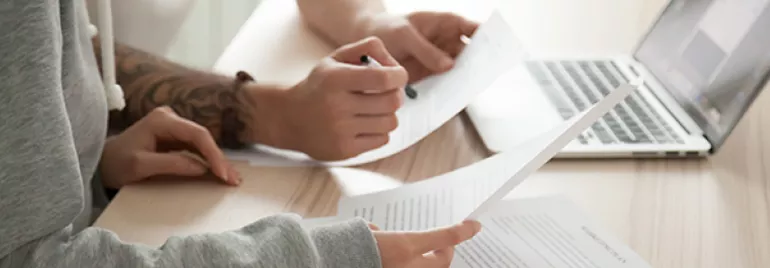 Two people reviewing documents at a desk with a laptop.