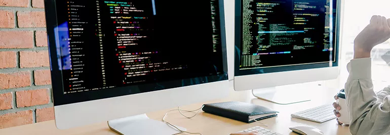 Two computer monitors displaying code in an office setting with two people working at a desk.