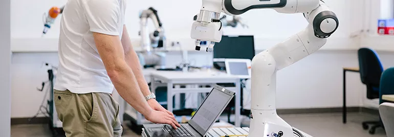 Engineer working on a laptop next to a robotic arm in a lab.
