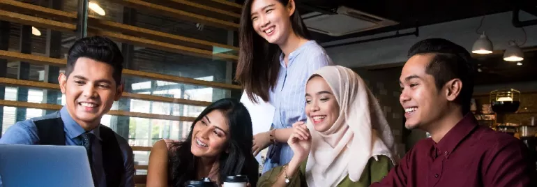 Five people gathered around a laptop, smiling and holding coffee, in an office setting.