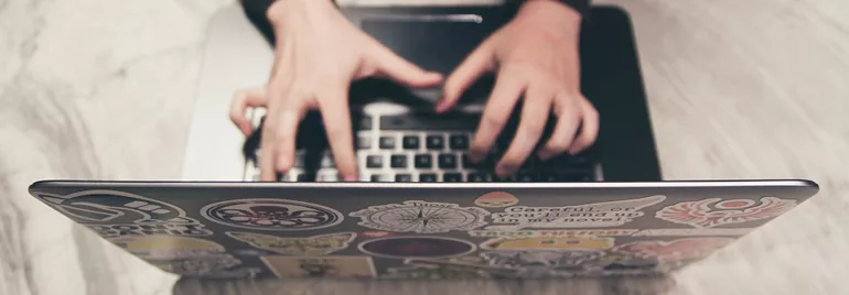 Hands quickly typing on a laptop covered with various colorful stickers.