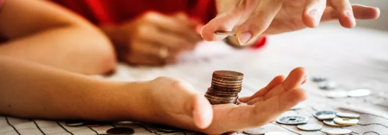 Hand holding a stack of coins, with another hand adding a coin to the stack on a table.