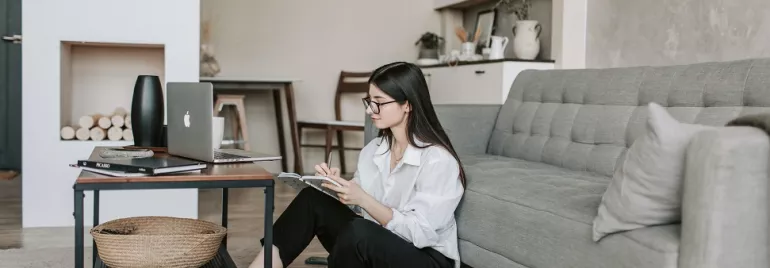 Woman sitting on floor near sofa, reading a book next to a laptop.