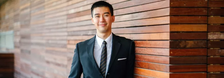 Man in a suit standing by a wooden wall, smiling.