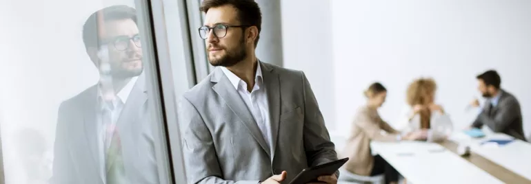 Businessman in a suit holding a tablet and looking out a window, with colleagues working at a table in the background.