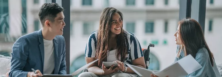 Three people in a meeting, smiling and discussing with papers and tablets.
