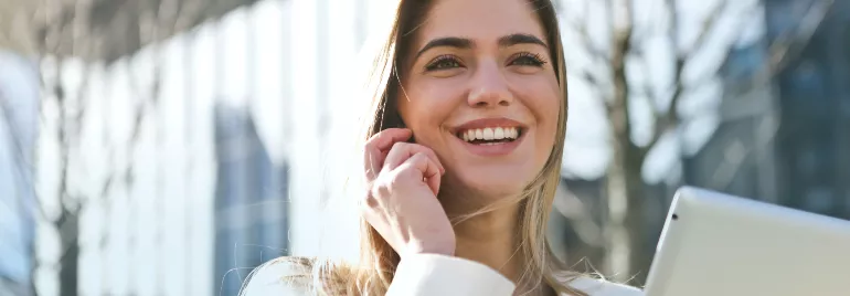 A Caucasian woman smiling with teeth while holding a mobile phone to her right ear, wearing a long-sleeved white top, in an outdoor setting with a building and some trees behind her.