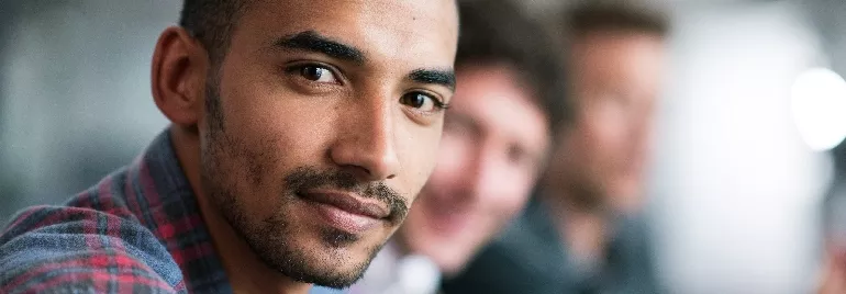 Man in a plaid shirt smiling with blurred colleagues in the background.