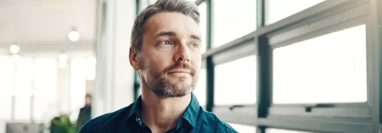 Man with a beard looking out a large window, wearing a blue shirt.