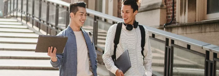 Two students with laptops walking and talking outside.