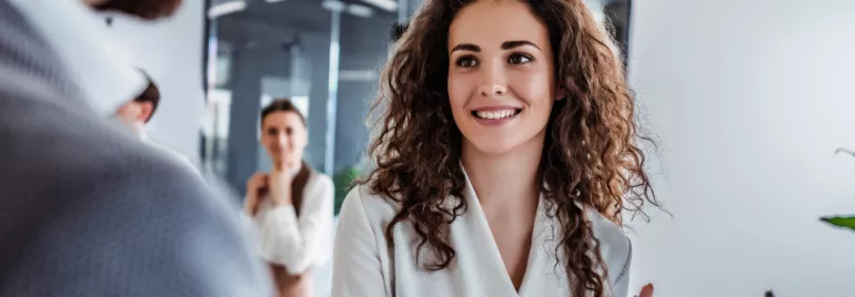 Person with curly hair smiling in office setting, two colleagues in the background.